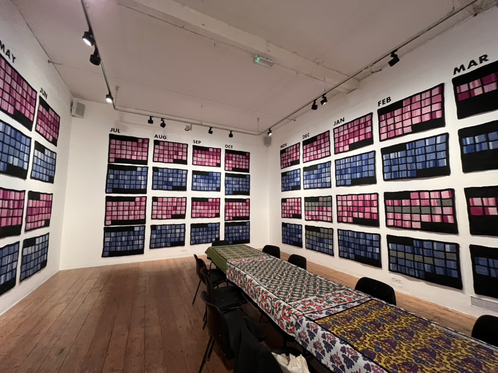 A gallery room, with fabrics containing knitted pink and blue squares, arranged in a calendar. A table with chairs sits in the middle of the room.